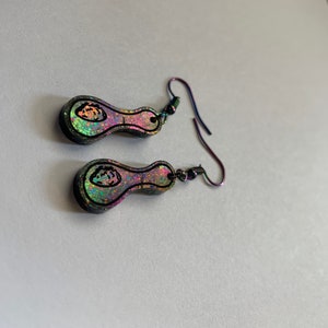 Może przedstawiać: Para iryzujących kolczyków w kształcie łyżki z czarnymi konturami i haczykami na uszy. Kolczyki mają kolorową, mieniącą się powierzchnię z czarnym wzorem pośrodku. Haczyki są w ciemnym, metalicznym kolorze.