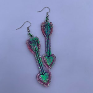 May include: A pair of iridescent pink and green arrow earrings with glitter accents. The arrowheads are shaped like hearts.