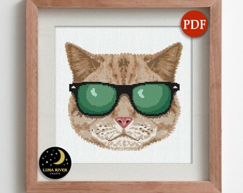 May include: Cross-stitch pattern of a cat wearing black sunglasses with green lenses. The cat has tan and brown fur, a pink nose, and is framed in a wooden frame. The image includes a "PDF" icon and a "Luna River Crafts" logo.