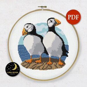 May include: Cross stitch pattern featuring two puffins standing on a rock with a blue ocean background. The pattern is in a round embroidery hoop.