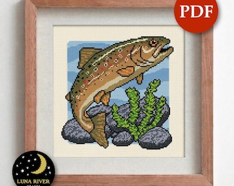 May include: A cross-stitch pattern PDF featuring a brown trout jumping over dark grey rocks and green aquatic plants against a light blue background. The design is displayed within a wooden frame, with a red PDF circle icon in the top right corner.
