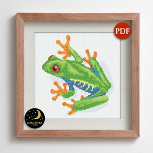 May include: Cross stitch pattern featuring a green and blue frog with orange feet and red eyes. The pattern is in a wooden frame with a white background. The pattern is labeled "PDF" and "Luna River Crafts".