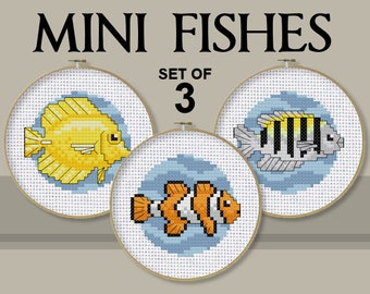 May include: A set of three circular embroidery hoops featuring cross-stitch patterns of a yellow fish, a black and white striped fish, and an orange and white clownfish on white fabric. The text MINI FISHES SET OF 3 is printed above the hoops.