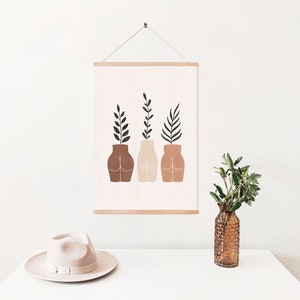 May include: A minimalist art print featuring three stylized figures with plants growing out of their heads. The figures are brown, beige, and terracotta, and the plants are green. The print is framed in a light wood frame and hanging on a white wall.