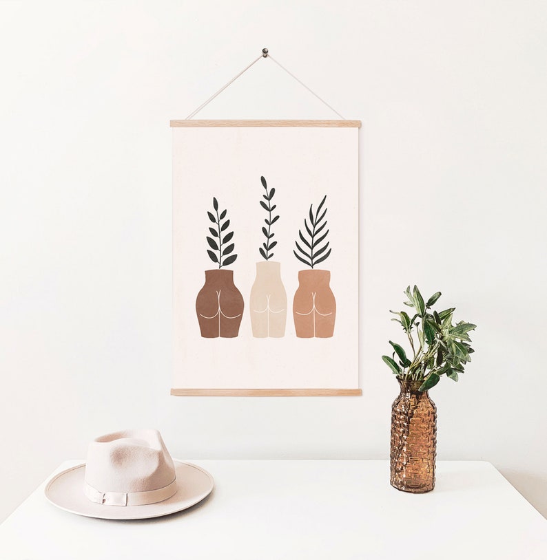 May include: A minimalist art print featuring three stylized figures with plants growing out of their heads. The figures are brown, beige, and terracotta, and the plants are green. The print is framed in a light wood frame and hanging on a white wall.