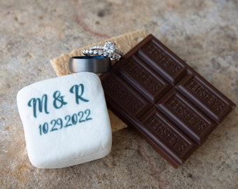May include: A white marshmallow with the text "M & R 10.29.2022" printed on it, a chocolate bar with the text "HERSHEY'S" printed on it, and two wedding rings on a graham cracker.