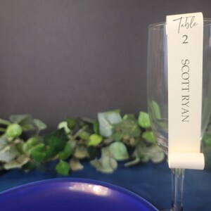 May include: A white table number card with the text "Table 2 SCOTT RYAN" attached to a clear glass champagne flute.