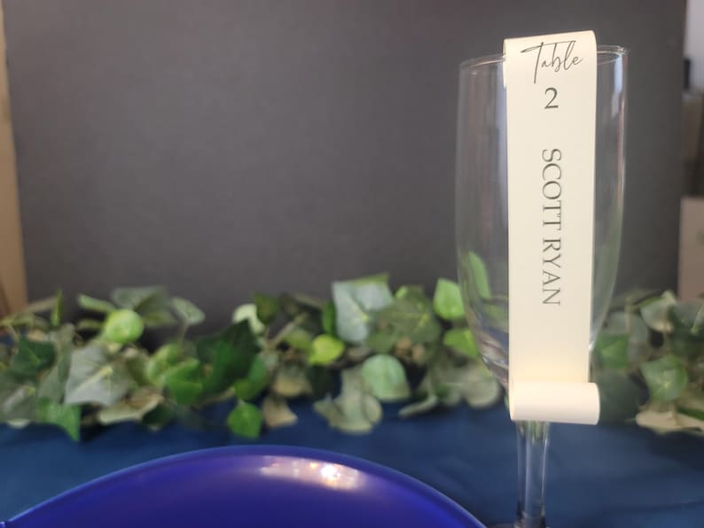 May include: A white table number card with the text "Table 2 SCOTT RYAN" attached to a clear glass champagne flute.