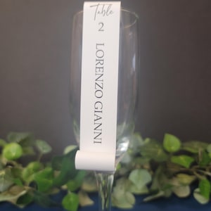 May include: White table number card with black text. The card says "Table 2 LORENZO GIANNI". The card is attached to a clear glass champagne flute.
