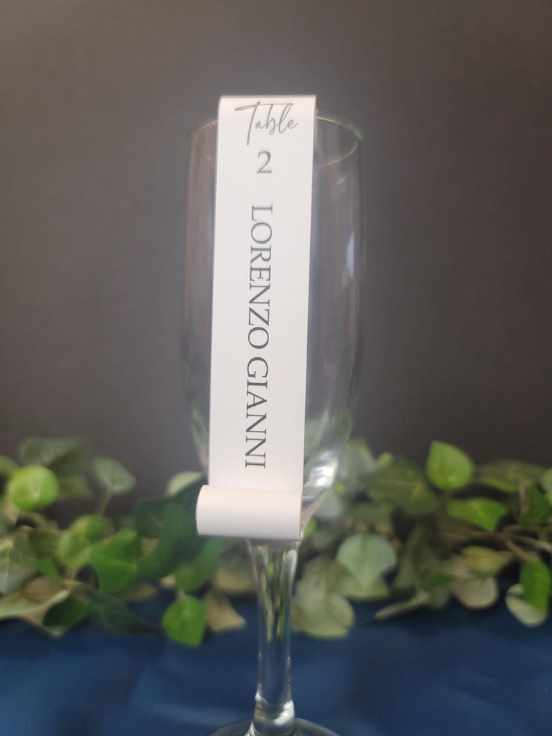 May include: White table number card with black text. The card says "Table 2 LORENZO GIANNI". The card is attached to a clear glass champagne flute.