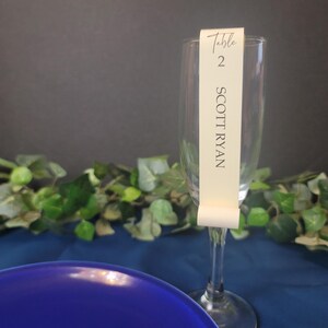 May include: A clear glass champagne flute with a white card attached to the stem. The card reads "Table 2 SCOTT RYAN".
