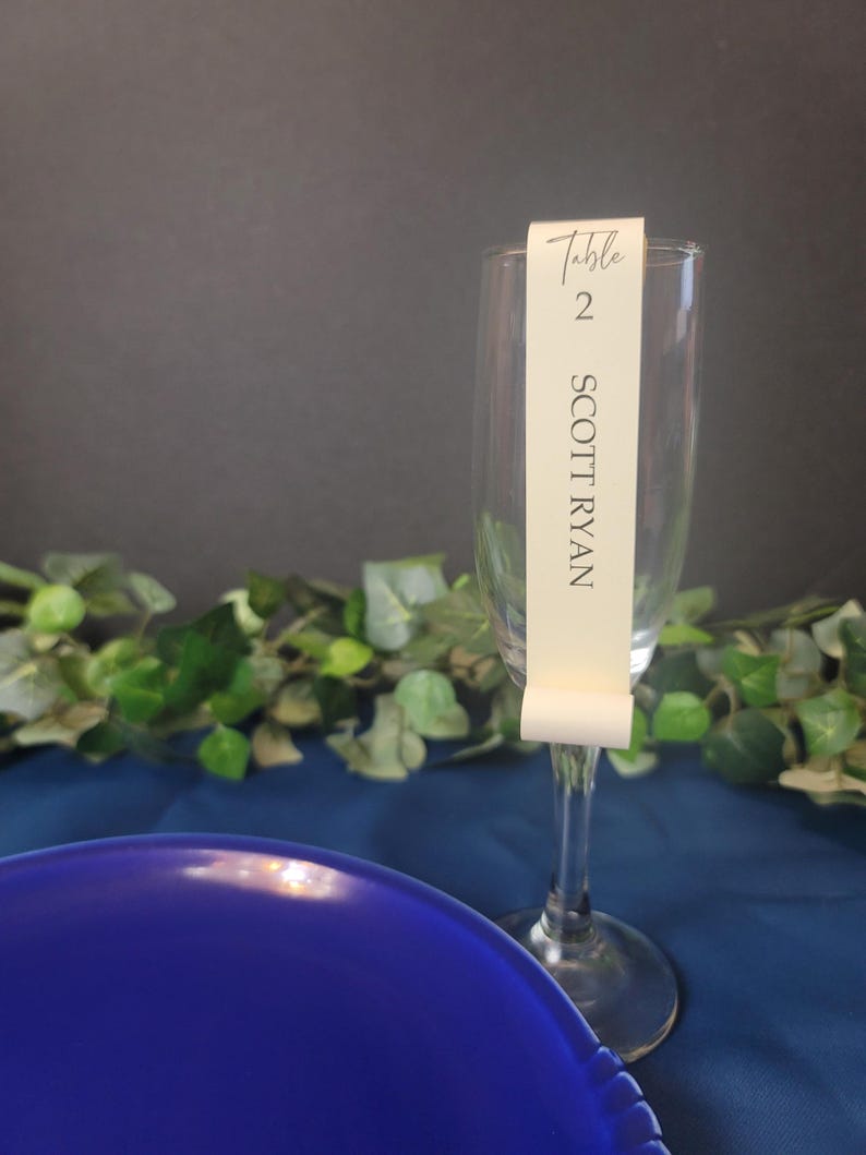 May include: A clear glass champagne flute with a white card attached to the stem. The card reads "Table 2 SCOTT RYAN".