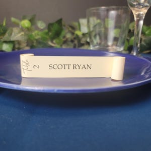 May include: A cream-colored table number card with the text "Table 2 SCOTT RYAN" printed in black. The card is rolled up and tied with a ribbon.
