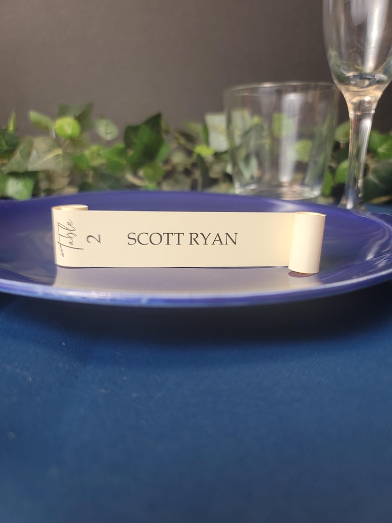 May include: A cream-colored table number card with the text "Table 2 SCOTT RYAN" printed in black. The card is rolled up and tied with a ribbon.