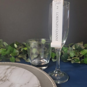 May include: A white table number card with black text that reads "Table 2 LORENZO GIANNI" is attached to a clear champagne flute. The flute is on a blue surface with a silver charger and a white marble plate.