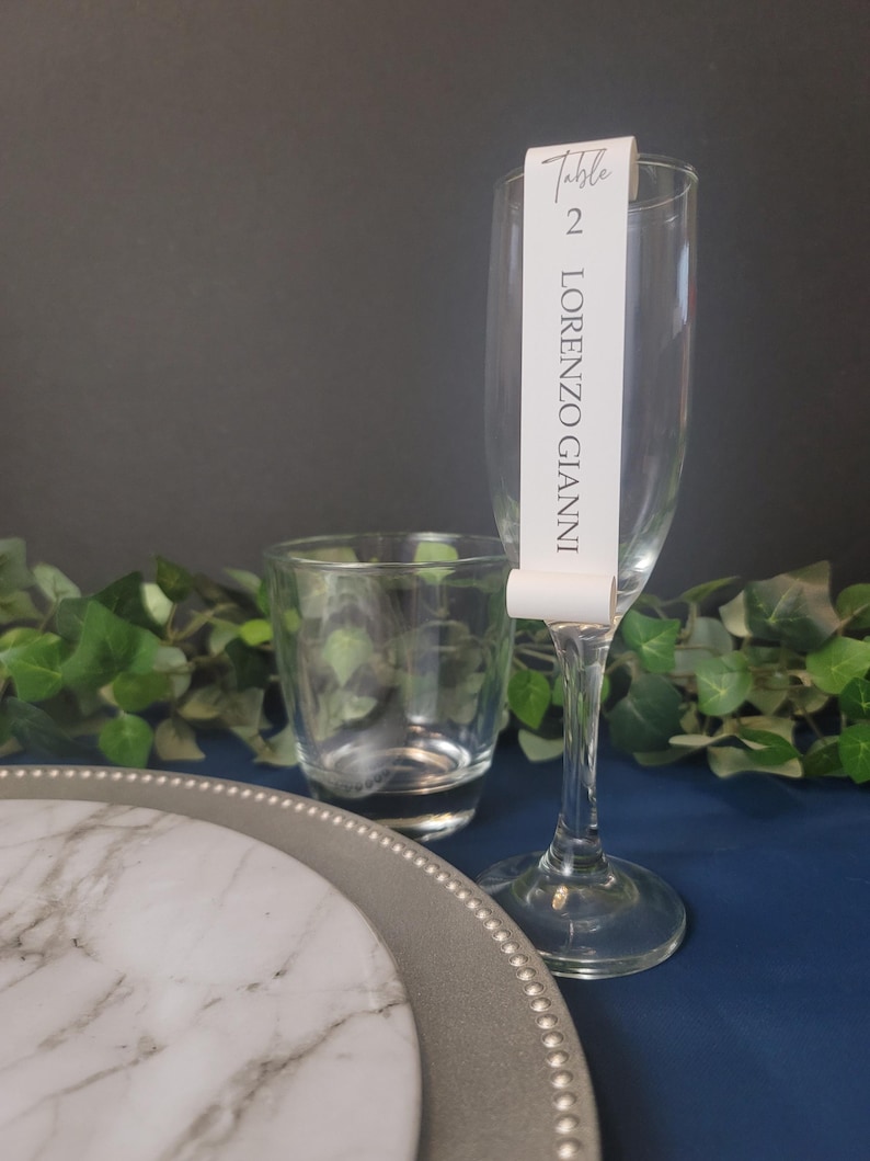 May include: A white table number card with black text that reads "Table 2 LORENZO GIANNI" is attached to a clear champagne flute. The flute is on a blue surface with a silver charger and a white marble plate.
