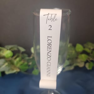 May include: White table number card with black text. The card is attached to a clear glass champagne flute. The card reads "Table 2 LORENZO GIANNI".