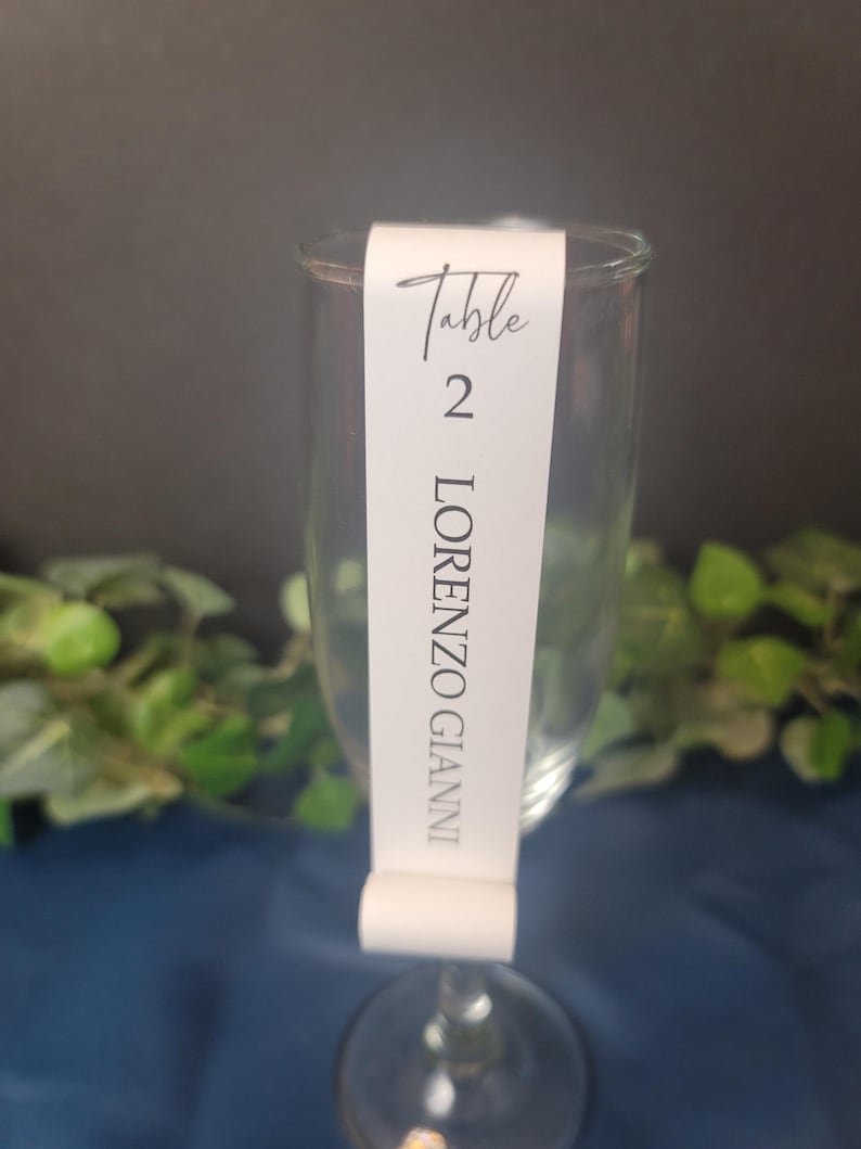 May include: White table number card with black text. The card is attached to a clear glass champagne flute. The card reads "Table 2 LORENZO GIANNI".