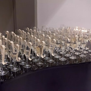 May include: A black grand piano with a row of champagne flutes arranged on top. Each flute has a small, rolled paper tag attached to the stem.