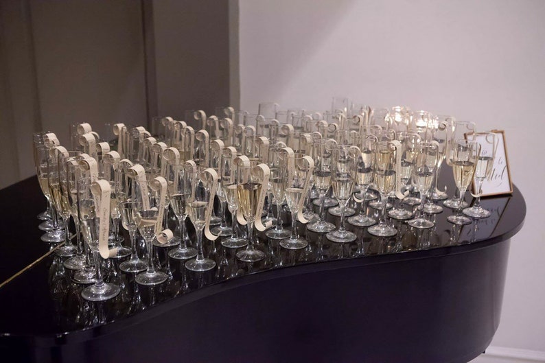 May include: A black grand piano with a row of champagne flutes arranged on top. Each flute has a small, rolled paper tag attached to the stem.