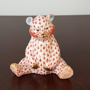May include: A small, red and white porcelain bear figurine with gold accents. The bear is sitting with its paws outstretched and has a detailed pattern of small red triangles on its body.