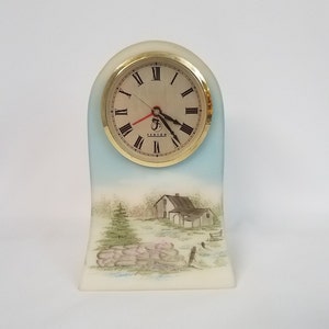 May include: A white ceramic clock with a gold rim and a painted scene of a house and trees. The clock face has Roman numerals and the word "Fenton" printed on it.
