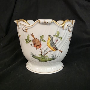 Peut inclure: Pot de fleurs en céramique blanche avec un bord festonné et une garniture dorée. Le pot présente un motif peint de deux oiseaux perchés sur une branche avec des feuilles vertes. La base est de style piédestal.