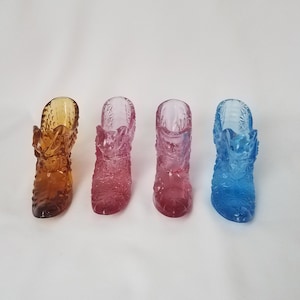 May include: Four glass boot-shaped vases in different colours: amber, pink, light pink, and blue. The vases are all decorated with a raised pattern.