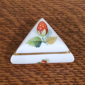 May include: A white, triangular porcelain trinket box with a hand-painted strawberry design. The lid features a red strawberry with green leaves. The box has a gold trim and a small strawberry painted on the side.