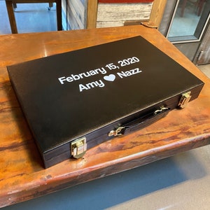 May include: A black leather game case with a gold latch and handle. The case is engraved with the text "February 15, 2020 Amy ♥ Nazz".