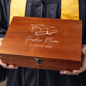May include: A wooden box with a graduation cap and diploma design, engraved with the name "Franklin Moore" and "CLASS OF 2026". The box is a warm brown color and has a metal clasp.