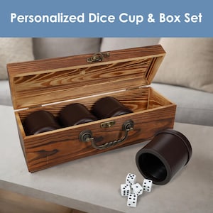 May include: A wooden box set with three brown dice cups inside and one cup outside. The box has a handle and clasp. Five white dice with black dots are next to the cup. The text "Personalized Dice Cup & Box Set" is at the top.