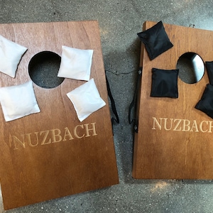 May include: Two wooden cornhole boards with the name "Nuzbach" engraved on them. One board has four white bean bags, the other has four black bean bags. Each board has a circular hole near the top.