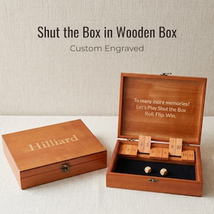 May include: A wooden Shut the Box game set featuring two boxes, one closed with the name Hilliard engraved on the lid, and one open revealing numbered tiles from 1 to 12, two dice, and the text To many more memories! Let's Play Shut the Box Roll. Flip. Win.