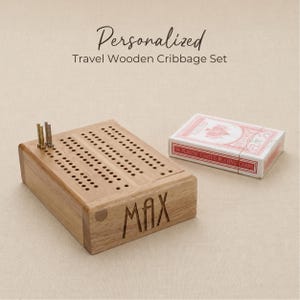 May include: A light brown wooden cribbage board with a grid of holes and the name MAX engraved on the front side. Two metal pegs are inserted into the board. Next to the board is a white box of plastic-coated playing cards. Text reads: Personalized Travel Wooden Cribbage Set.
