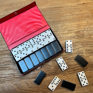 May include: A set of dominoes in a red case with black and white tiles. The dominoes are arranged in a rectangular shape with the black tiles on the bottom and the white tiles on top. The dominoes are scattered around the case on a wooden surface.