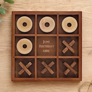 May include: A square wooden tic-tac-toe game board with a dark brown finish. The center square is engraved with the text June Birthday Girl. The board contains four light-colored wooden circular O pieces and five dark wooden X pieces.