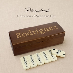May include: A rectangular dark brown wooden box with the name Rodriguez engraved on the lid. Beside the box are several off-white dominoes, each featuring the name Rodriguez printed in black ink, with one domino showing black pips.