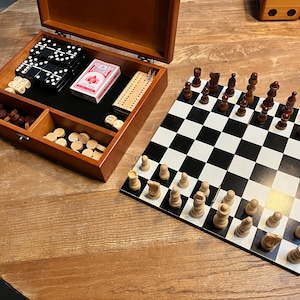 May include: A wooden box with a variety of games inside, including dominoes, dice, checkers, and a deck of cards. The box is open and the games are visible. There is also a chessboard with chess pieces on it.
