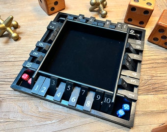 May include: A black Shut the Box dice game with numbered tiles and a central playing area. The game includes red and blue dice, and is surrounded by wooden dice and gold jacks.
