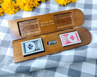 May include: Wooden cribbage board with two decks of playing cards. The board is open, showing the pegging board and card storage. The text "Rachel Ann May 24, 2024" is engraved on the board. The playing cards are red and blue.