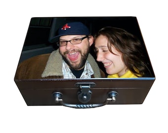 May include: A brown, vintage-style suitcase with a photograph on the lid. The photo features a smiling man wearing glasses and a baseball cap, and a smiling woman. The suitcase has a metal handle and clasp.