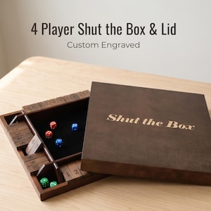 May include: A dark brown wooden 4-player Shut the Box game board with a matching lid. The lid features the engraved text Shut the Box. The open board has numbered tiles from 1 to 10 on four sides, a black felt center, and red, blue, and green dice.