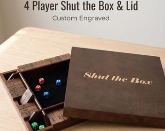 May include: A dark brown wooden 4-player Shut the Box game board with a matching lid. The lid features the engraved text Shut the Box. The open board has numbered tiles from 1 to 10 on four sides, a black felt center, and red, blue, and green dice.