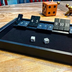May include: A black wooden game tray with a black felt lining. The tray has a row of numbered tiles from 1 to 12. Two white dice are on the felt.