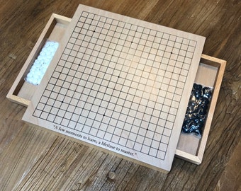 May include: A wooden Go board with a drawer on the side. The drawer contains black and white Go stones. The board has a quote on it that reads "A few moments to learn, a lifetime to master."