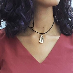 Puede incluir: Un collar con un colgante de oro con una pequeña figura detallada de una persona con una capa. El collar está en un choker negro.