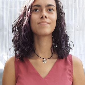 May include: A black cord necklace with a small, round pendant. The pendant has a silver-colored border and a star design with yellow and gold accents. The person in the image is wearing a red-pink top.