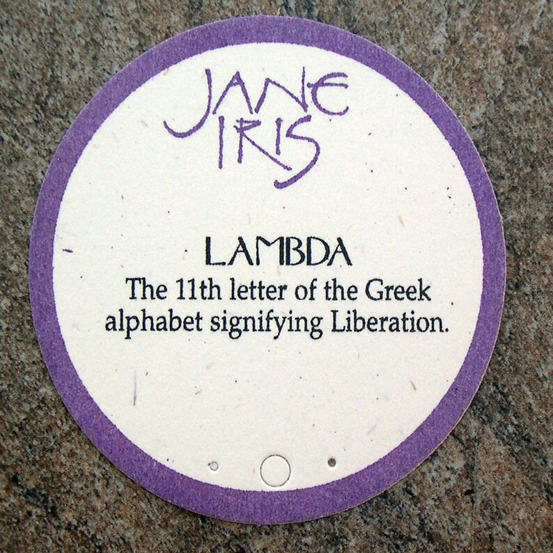 May include: A round, white card with a purple border. The card has the text "JANE IRIS" at the top and "LAMBDA" in a larger font below. The text "The 11th letter of the Greek alphabet signifying Liberation." is printed below the word "LAMBDA".