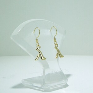 Puede incluir: Un par de pendientes de aro dorados con un diseño abstracto simple. Los pendientes están en un expositor de plástico transparente.